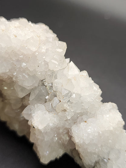 A close-up view of a cluster of sparkly white quartz crystals from The Crystalary's AUCTION Quartz over Fluorite- unknown Chinese Locality, against a dark background. The crystals are densely packed, with many small, sharp points catching the light and creating a sparkling effect. The varied, irregular surfaces highlight the natural formation of this mineral from its Chinese locality.