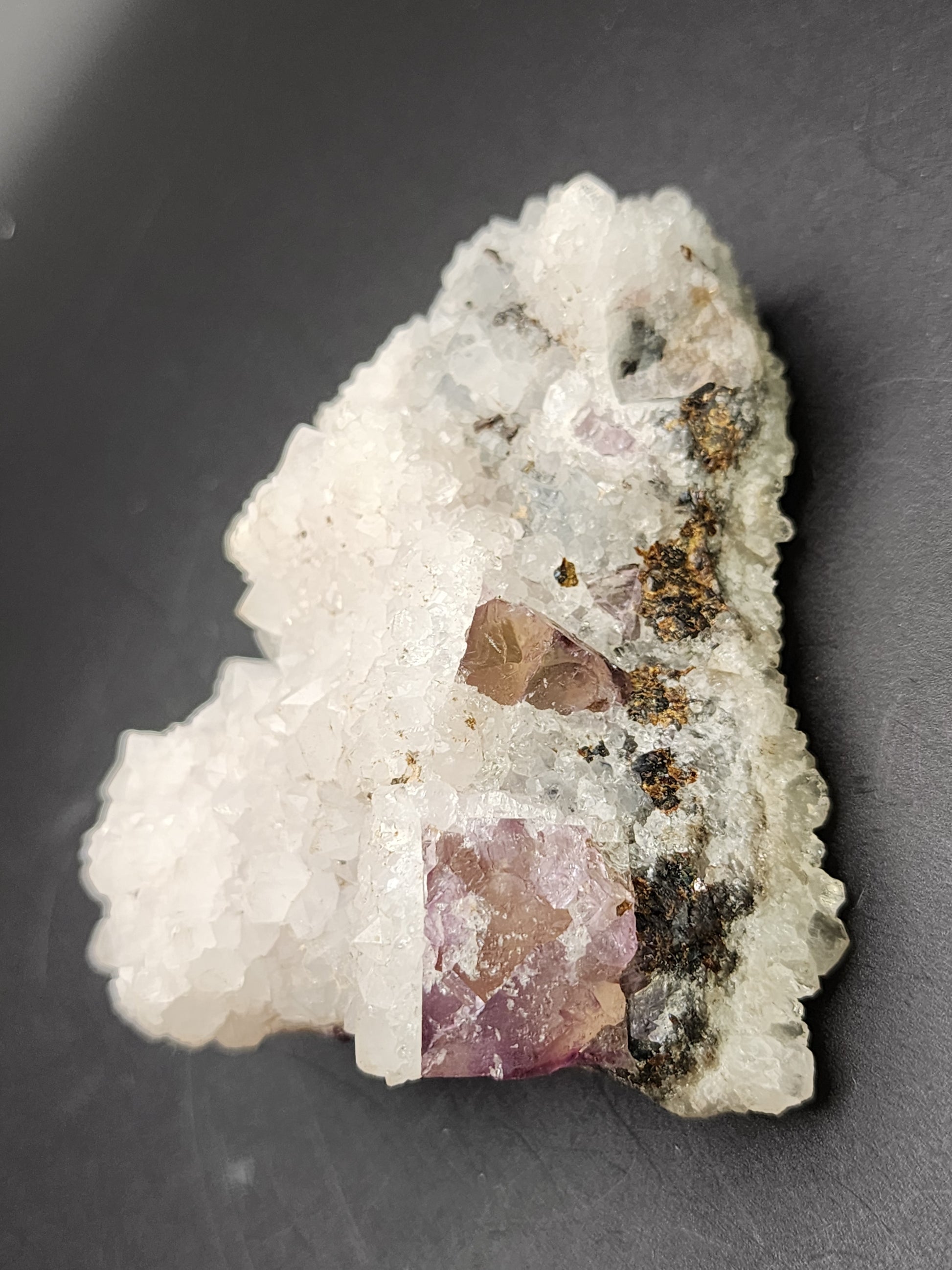 A close-up view of AUCTION Quartz over Fluorite from an unknown Chinese locality, offered by The Crystalary. The sparkly white quartz showcases translucent crystals adorned with purple and brown inclusions, set against a black background. The rough and irregular surface highlights the natural formation of these captivating minerals.