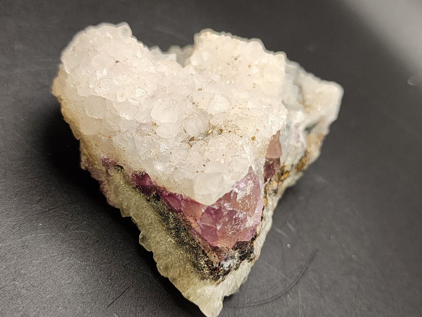 A close-up image showcases The Crystalary's AUCTION Quartz over Fluorite from an unknown Chinese locality. The raw mineral specimen features clusters of sparkling, translucent crystals on a light-colored base. The crystals exhibit a pale pink hue with some areas showing purple coloration, reminiscent of fluorite from China. The specimen is set against a plain dark background.