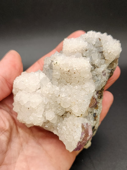 A hand holds the AUCTION Quartz over Fluorite from The Crystalary, a remarkable mineral specimen with small, sparkly white quartz formations densely packed over its surface. Set against a solid black background that accentuates the texture and structure of the mineral, this stunning piece originates from an unidentified locality in China.