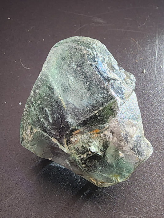 A rough, translucent mineral with a predominantly greenish hue and varying shades of color sits on a dark, textured surface. This AUCTION Fluorite from The Crystalary, sourced from the Superior Mine in Mambilla Plateau, Nigeria, has an uneven, jagged surface that catches the light, revealing reflective and glistening areas.