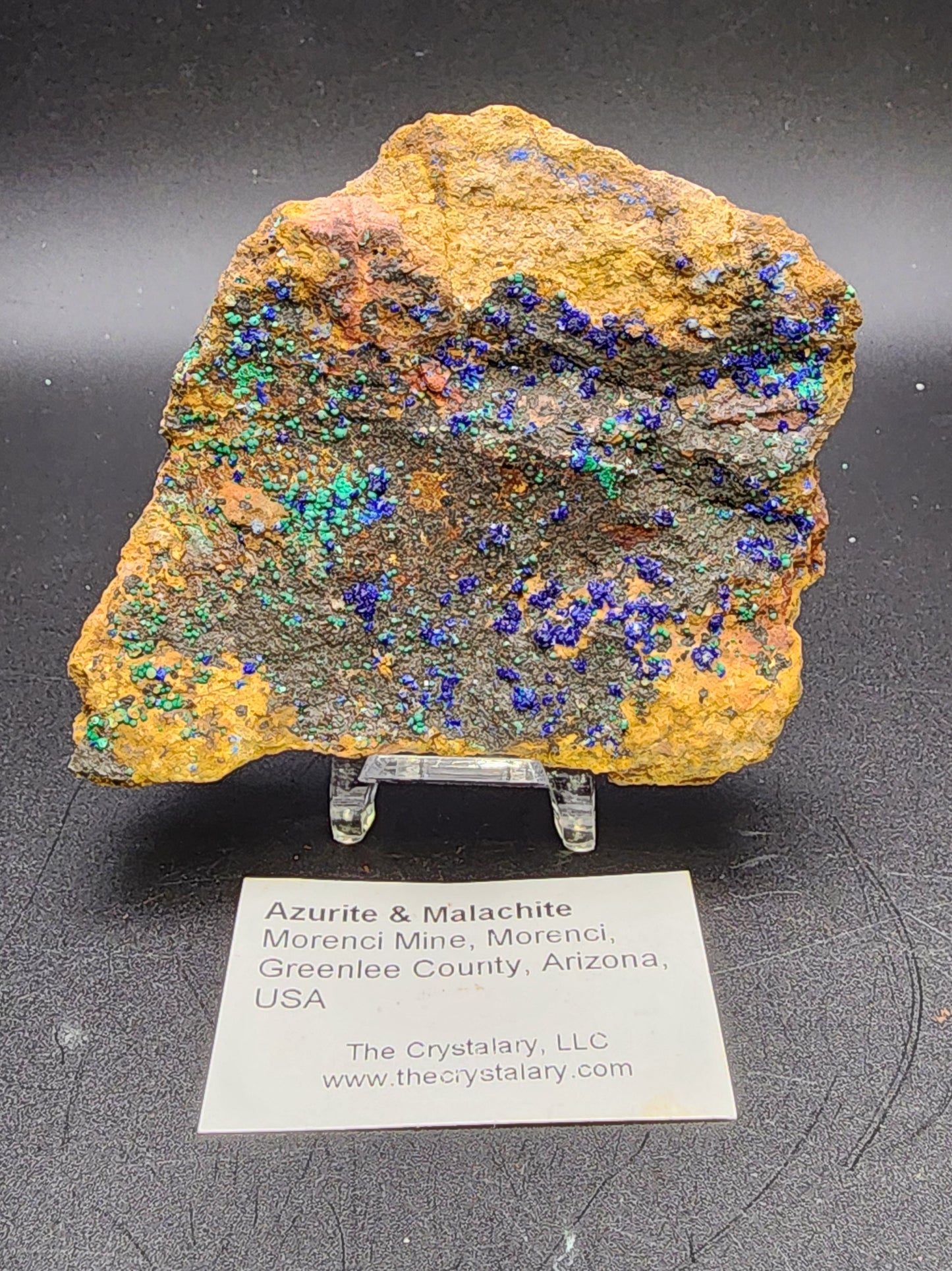 This AUCTON Azurite & Malachite rock specimen from the renowned Morenci Mine in Greenlee County, Arizona, USA features vibrant blue and green crystalline sections on a yellow-brown matrix. The display comes with a detailed label and can be found at The Crystalary website, thecrystalary.com.