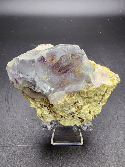 The AUCTION Fluorite from Yindu Mine, Hexigten Banner, Chifeng City, Inner Mongolia, China by The Crystalary is a large, rough crystalline mineral specimen featuring light purple coloration accented with blue-zoned fluorite sections. Resting on a beige rock matrix, the partially transparent crystal showcases a lustrous surface and is elegantly displayed on a clear acrylic stand against a dark background.