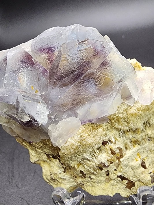 A large crystal formation of blue-zoned fluorite with translucent light purple and blue hues rests on a textured, pale yellow rock base. The crystal's surface appears smooth and slightly reflective, while the rock base remains rough and uneven. This stunning specimen, known as "AUCTION Fluorite" from The Crystalary, is sourced from the Yindu Mine in Hexigten Banner, Chifeng City, Inner Mongolia. The background features a simple dark gradient.