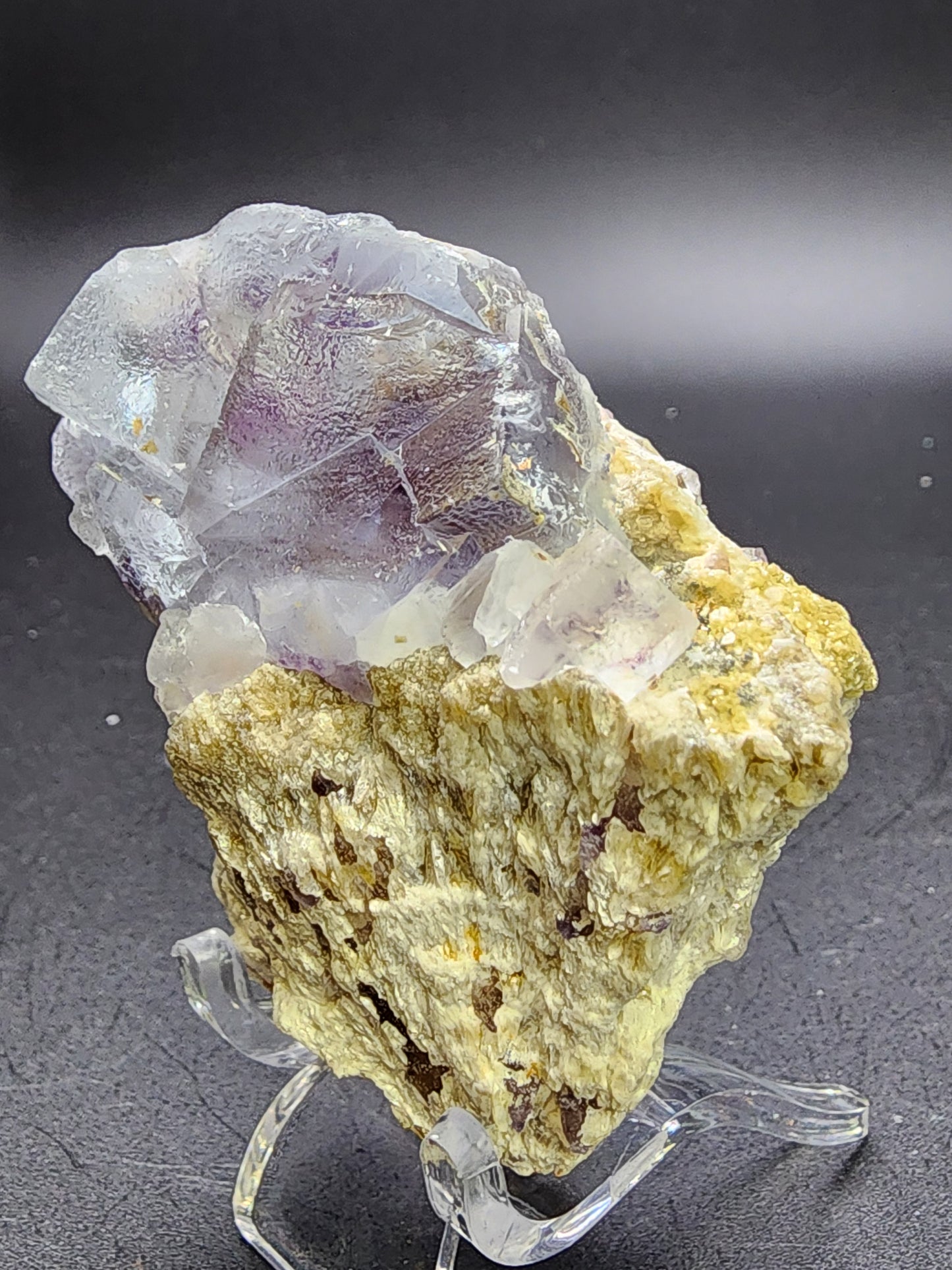 A pale purple AUCTION Fluorite crystal from the renowned Yindu Mine in Inner Mongolia, China, rests on a rough, yellowish-beige base. The translucent specimen is elegantly displayed on a transparent stand against a dark backdrop. This exquisite piece comes from The Crystalary and hails specifically from Hexigten Banner, Chifeng City.