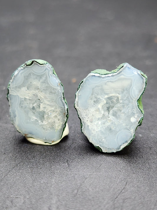 A close-up of two halves of "AUCTION- Tabasco Geodes- Tabasco, Mexico" from The Crystalary, showcasing a light blue geode with greenish edges. The geode reveals a crystalline interior with intricate patterns and textures, reflecting light and displaying varying shades of blue and white. The background is a solid dark surface.