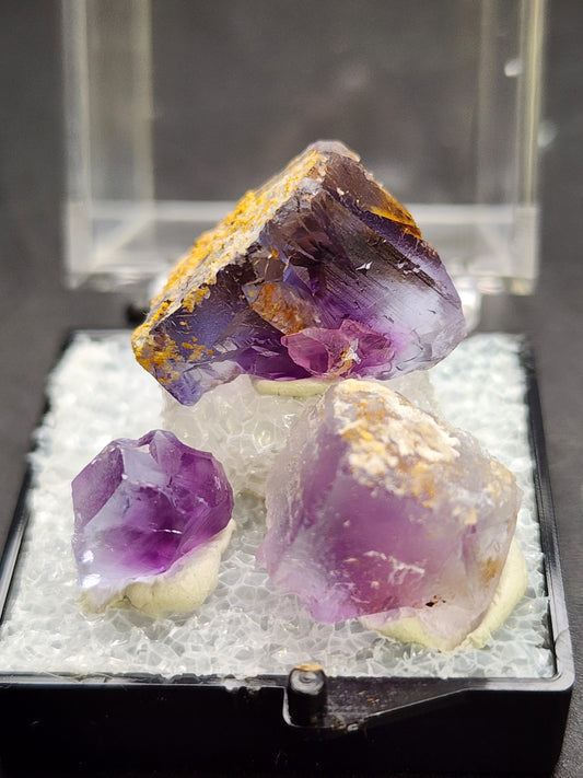 Three purple fluorite crystals of varying sizes, partially coated with a yellow-brown mineral, are displayed on a white, textured base within a clear display case. These exquisite specimens from The Crystalary's AUCTION Fluorite- Cow Poke Pocket collection originate from the Blanchard Mine in the Hansonburg District of Socorro County, New Mexico, and have garnered attention at the New Mexico Mineral Symposium.