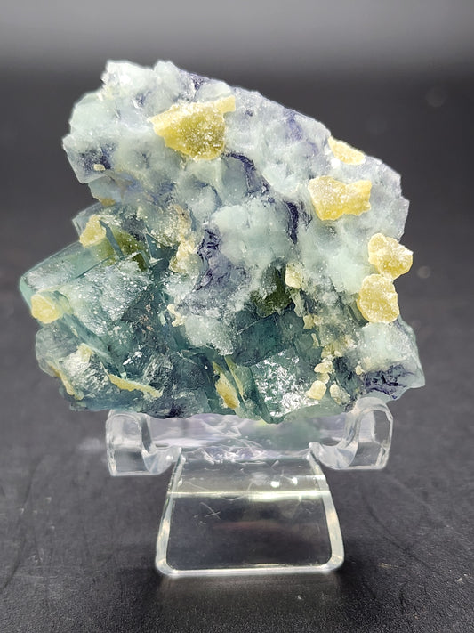 A cluster of translucent, multi-colored fluorite crystals from The Crystalary's collection, including green and purple specimens from the Yindu Mine in Hexigten Banner, Chifeng City, Inner Mongolia, China, rests on a small clear stand. The crystals exhibit shades of blue, green, and purple with some yellowish mineral deposits scattered across the surface. The background is a plain black surface.