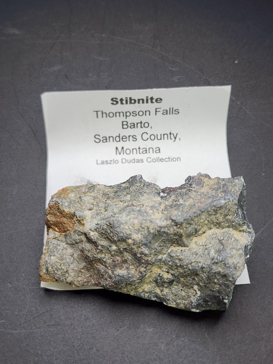 A stibnite specimen from The Crystalary rests on a dark surface with a label behind it reading: "AUCTION Stibnite- Thompson Falls, Barto, Sanders County, Montana, USA." The stibnite has a rough, grayish appearance with some brownish discoloration.