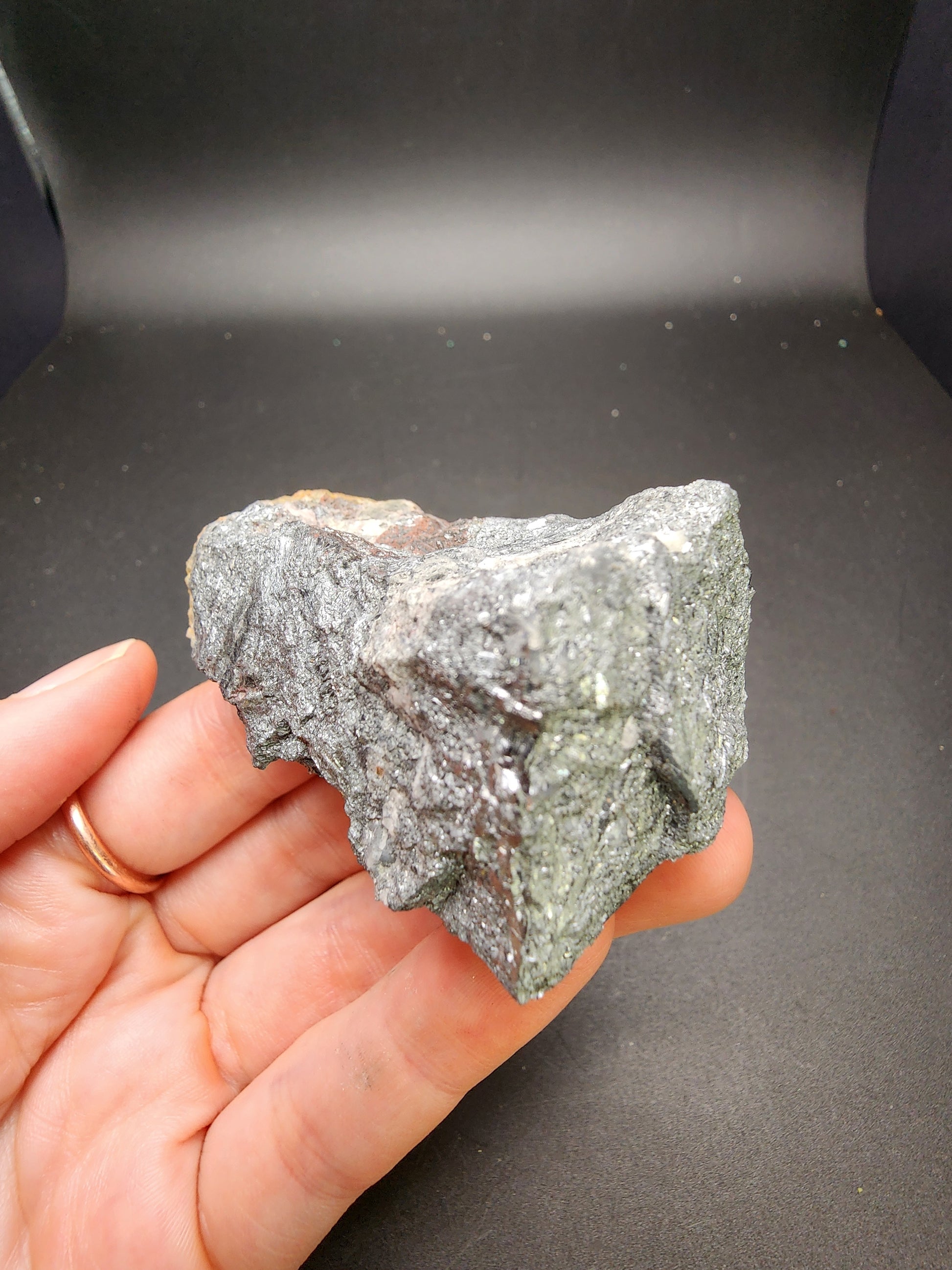 A hand adorned with a gold ring on the ring finger is holding a rough, metallic-looking specimen of AUCTION Stibnite from Thompson Falls, Barto, Sanders County, Montana, USA against a dark background. The mineral, featured as part of The Crystalary's Laszlo Dudas Collection, boasts a glistening surface characterized by a mix of dark silver and grayish-green hues.