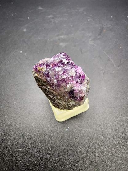 A rough, unpolished amethyst crystal with a mix of deep purple and white hues sits on a small, yellowish base against a dark background. Nearby, "AUCTION Fluorite- Annabel Lee Mine & Minerva No. 1 Mine, Hardin County, Illinois, USA" by The Crystalary adds an intriguing burst of color. The crystal's jagged texture and natural formation are clearly visible.