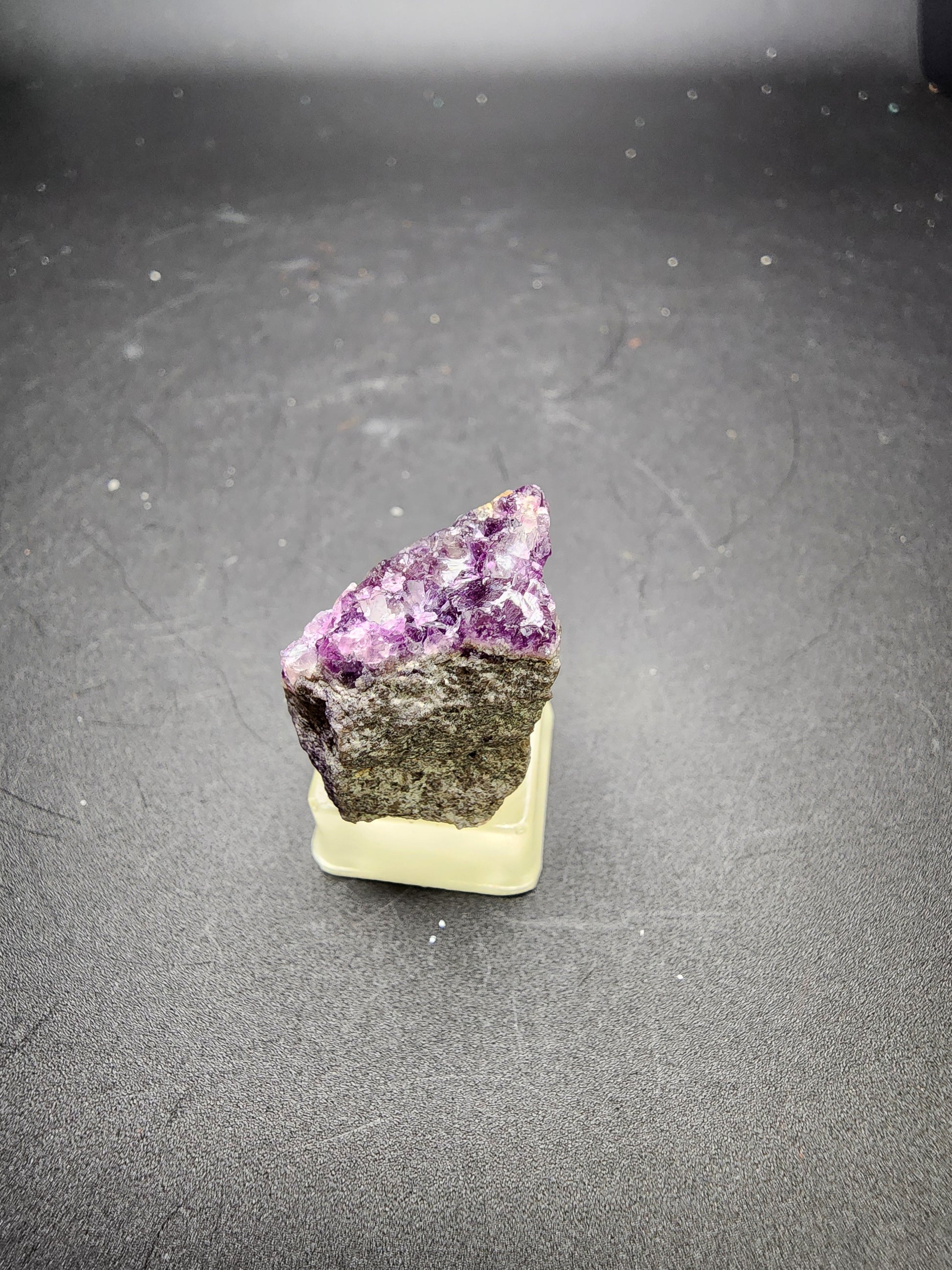 A rough mineral specimen from The Crystalary’s AUCTION Fluorite collection, originating from the Annabel Lee Mine and Minerva No. 1 Mine in Hardin County, Illinois, features purple and white crystalline structures atop a gray base, displayed on a small yellow stand against a black background.