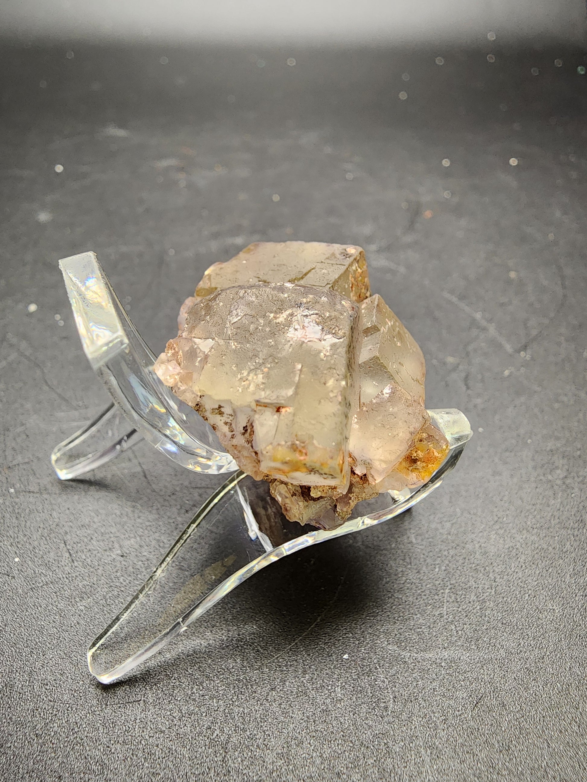 An AUCTION Fluorite specimen from the El Hammam Mine in Ait Mimoune Caïdat, Khémisset Cercle, Khémisset Province, Rabat-Salé-Kénitra Region, Morocco by The Crystalary features translucent, cubic crystals perched on a clear plastic stand against a black background. This fluorite cluster displays a slight hint of color and natural imperfections, showcasing the rugged beauty of the mineral.