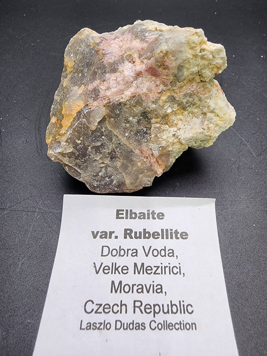 A rough, uncut crystal rock with shades of white, pink, and grey is displayed on a black surface. A label beneath it reads: "AUCTION Elbaite var. Rubellite- Dobra Voda, Velke Mezirici, Moravia, Czech Republic," proudly part of the Lazlo Dudas Collection by The Crystalary.