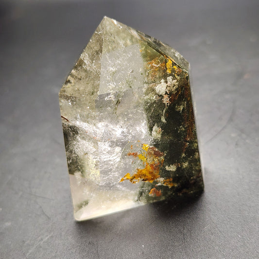 A transparent polished crystal from The Crystalary, identified as AUCTION - Garden Quartz Point, stands upright against a dark background. The inclusions within the crystal are reminiscent of Garden Quartz and vary in color, including patches of green and yellow, giving it a distinct and intricate appearance.