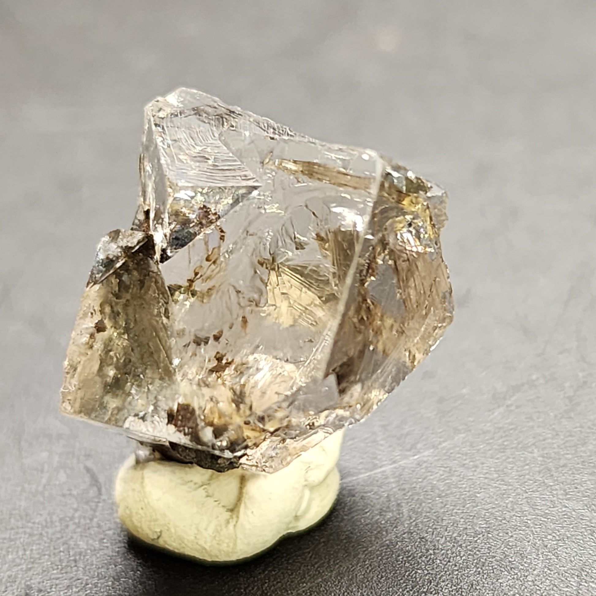 A close-up image of a clear, roughly rectangular crystal with jagged edges, identified as "AUCTION Fluorite- Bull Vein" from the Lady Annabella Mine in Eastgate, Weardale, Co. Durham, England. This exquisite piece by The Crystalary is mounted on a small piece of putty and placed on a dark, smooth surface. Swirls and inclusions are visible within the crystal, giving it a textured appearance reminiscent of Bull Vein Miniatures from the same mine.