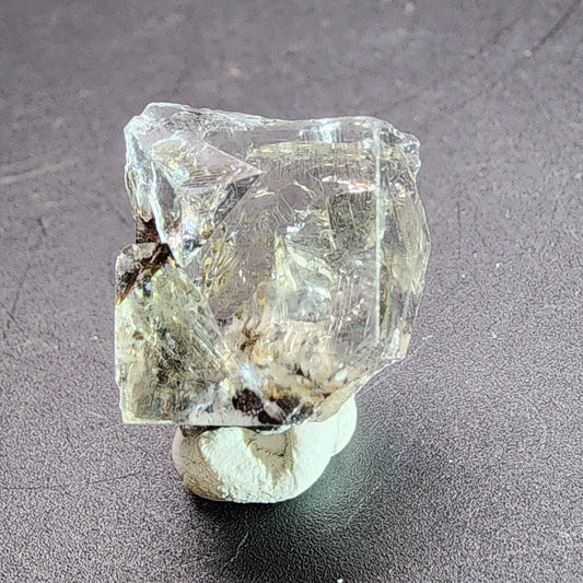 A detailed close-up photo features the rough, translucent AUCTION Fluorite gemstone with a pale, slightly yellow hue from The Crystalary. This irregularly shaped stone is positioned on a small mound of white putty against a dark surface to help it stand upright. Sourced from the Bull Vein at the Lady Annabella Mine in Eastgate, Weardale, Co. Durham, England, this unique gem is showcased effectively for display.