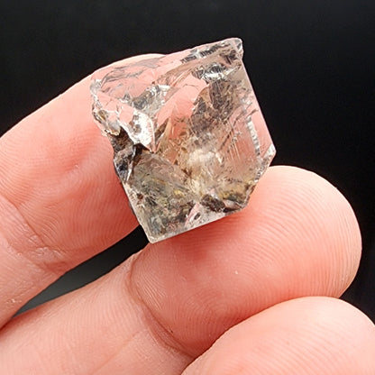 A close-up shot of a hand holding a transparent, roughly cut fluorite gemstone from The Crystalary's AUCTION Fluorite – Bull Vein collection, sourced from the Lady Annabella Mine in Eastgate, Weardale, Co. Durham, England. The person's fingers gently grip the mineral against a black background, highlighting its reflective surfaces and natural inclusions.