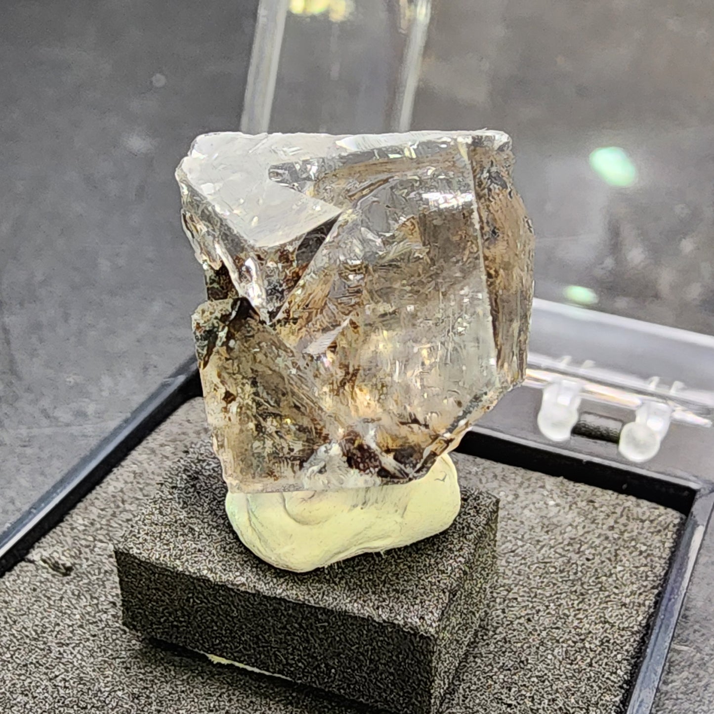 A close-up image of the transparent, geometric fluorite crystal from The Crystalary, mounted on a piece of foam in a display box. This particular piece is sourced from the Bull Vein, Lady Annabella Mine, Eastgate, Weardale, Co. Durham, England. The crystal has visible internal fractures and imperfections with a light brown tint in some areas. The background is blurred and dark.
