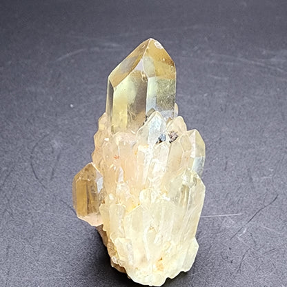 On a dark surface sits a mini points cluster of yellowish transparent quartz crystals with a prominent upright point. The Kundaline Citrine cathedral candle-like crystals from The Crystalary exhibit a rough texture and vary in size, forming an intricate, jagged structure that mirrors the unique beauty found in Lwena specimens from Katanga, Democratic Republic of Congo.