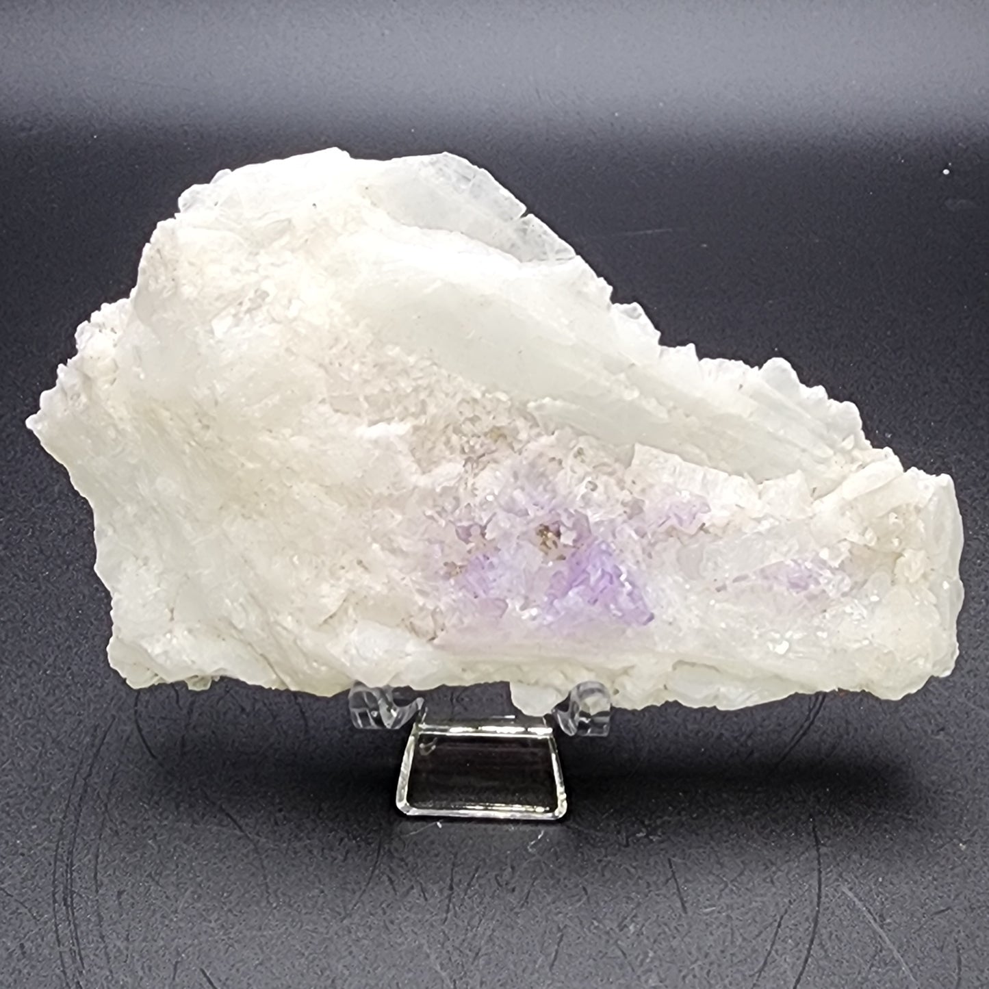 A large, white crystalline mineral specimen with hints of light purple tint at the center is displayed on a small, clear acrylic stand against a plain, dark background. The AUCTION Fluorite on Celestine from the Tule Mine in Melchor Múzquiz, Mun. de Melchor Múzquiz, Coahuila, Mexico by The Crystalary has a rough, jagged texture and irregular shape reminiscent of finds from this renowned location.