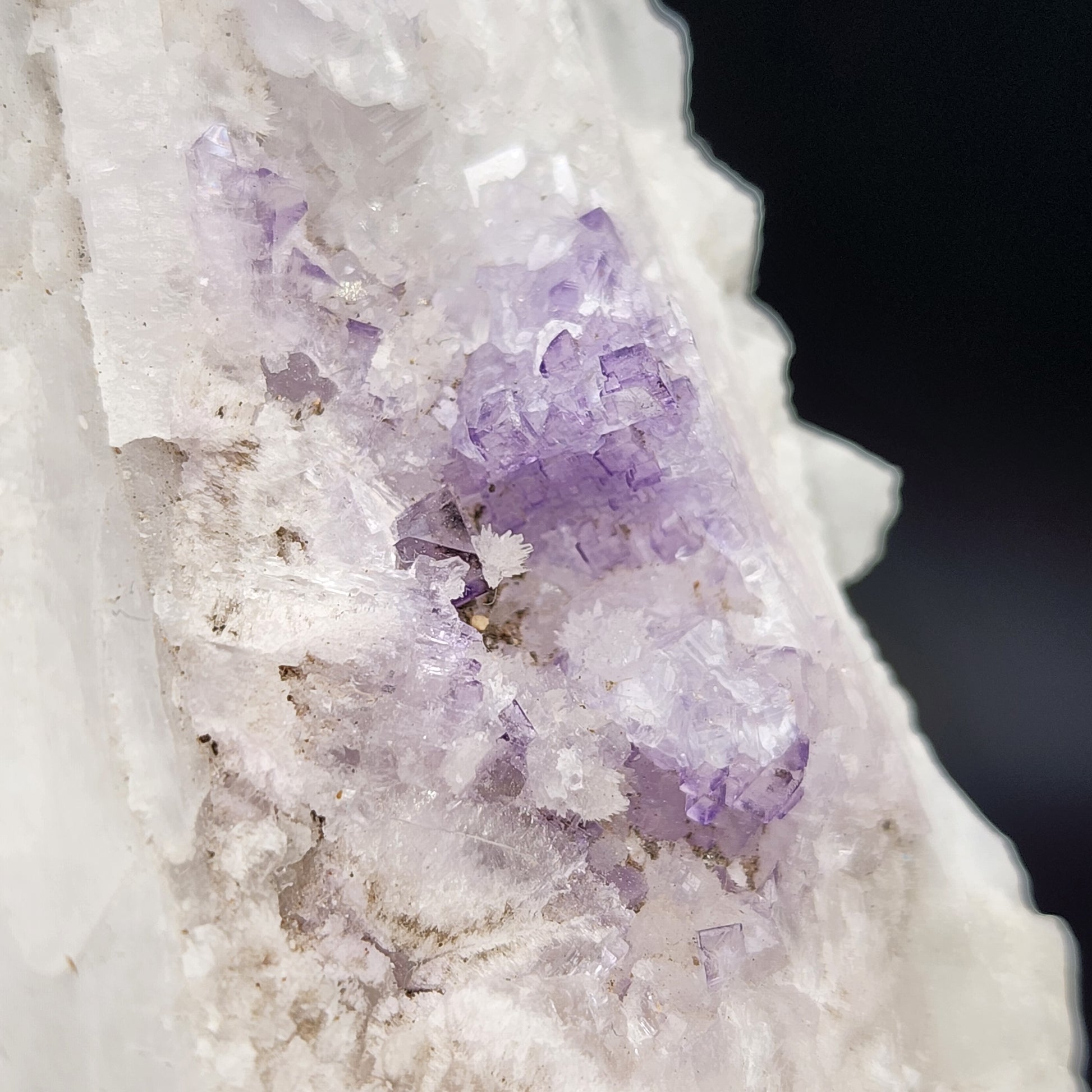 Close-up photo of AUCTION Fluorite on Celestine from The Crystalary, showcasing a rough crystal formation, primarily white with patches of translucent purple fluorite. The irregular surface features jagged edges and a mix of textures, highlighting the natural beauty of the mineral from the Tule Mine in Melchor Múzquiz, Coahuila, Mexico. The background is dark and out of focus.