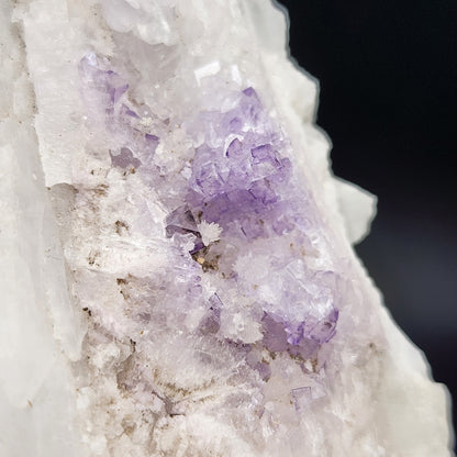 Close-up photo of AUCTION Fluorite on Celestine from The Crystalary, showcasing a rough crystal formation, primarily white with patches of translucent purple fluorite. The irregular surface features jagged edges and a mix of textures, highlighting the natural beauty of the mineral from the Tule Mine in Melchor Múzquiz, Coahuila, Mexico. The background is dark and out of focus.