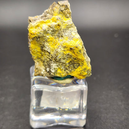 A rough mineral specimen, identified as AUCTON Pottsite, showcases bright yellow and earthy gray tones. It is elegantly displayed on a clear, square pedestal against a dark, blurred background. This rare mineral has been sourced from the renowned Linka Mine in the Spencer Hot Springs Mining District of Lander County, Nevada, USA and is offered by The Crystalary.