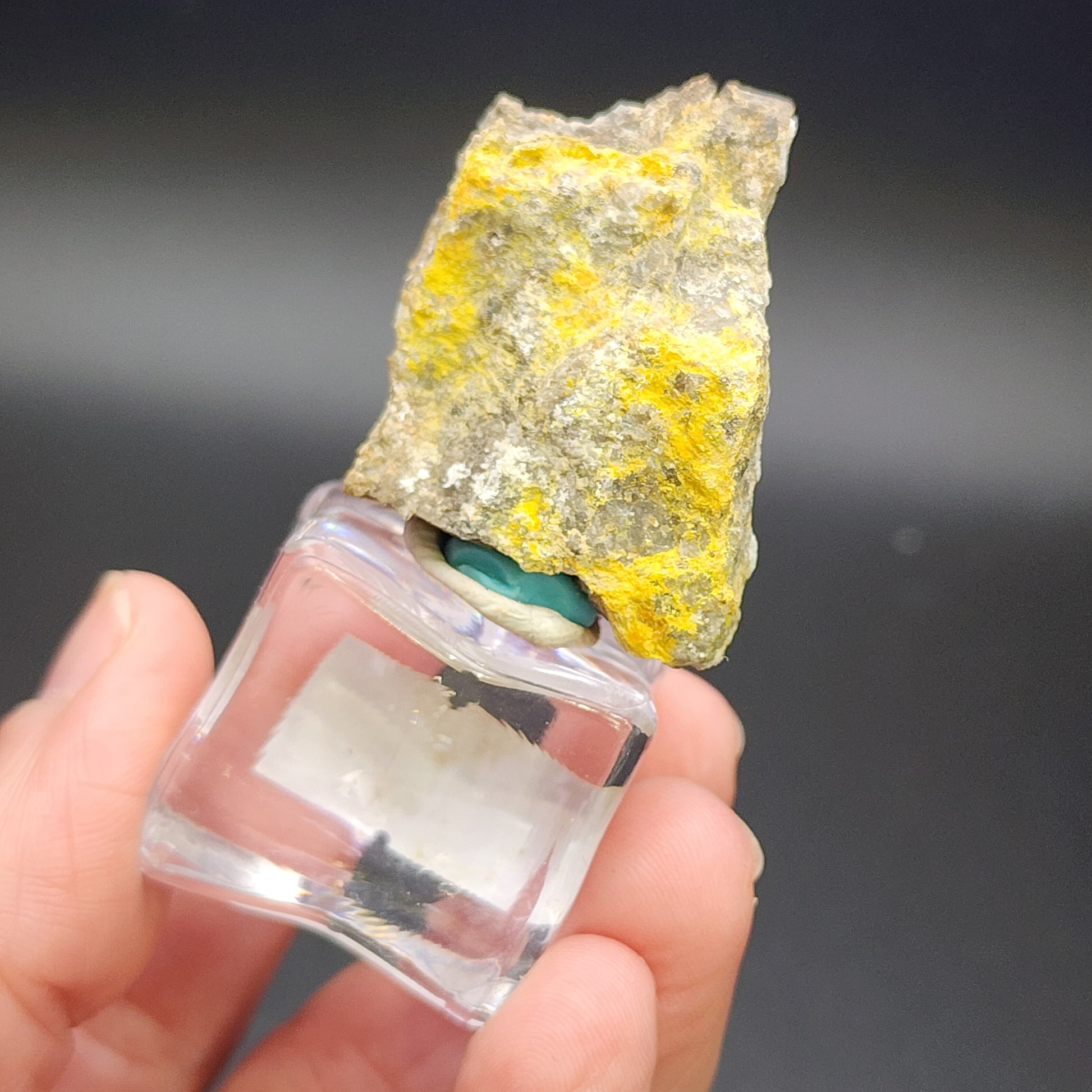 A hand holds a small transparent square container with a jagged, yellow-streaked rock placed on top. The rock, identified as AUCTON Pottsite from the Linka Mine, Spencer Hot Springs Mining District, Lander County, Nevada, USA by The Crystalary, has a rough texture and appears to be a rare mineral sample. The container is set against a black background.