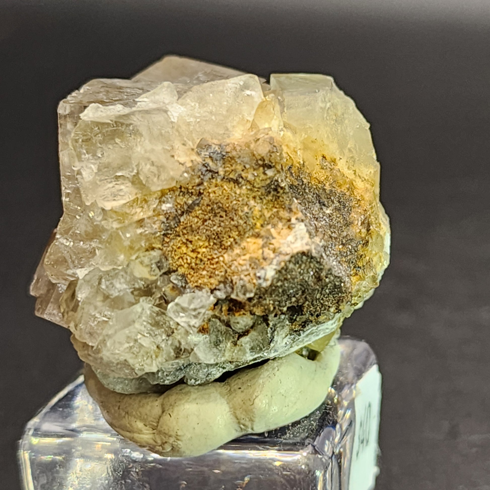 Displayed on a clear cubic stand against a dark background is the AUCTION Fluorite from The Crystalary. Hailing from the Bull Vein, Lady Annabella Mine in Eastgate, Weardale, Co. Durham, England, this translucent rough crystal features yellow and brown inclusions. The mounted cluster specimen boasts an irregular shape with sharp, jagged edges.