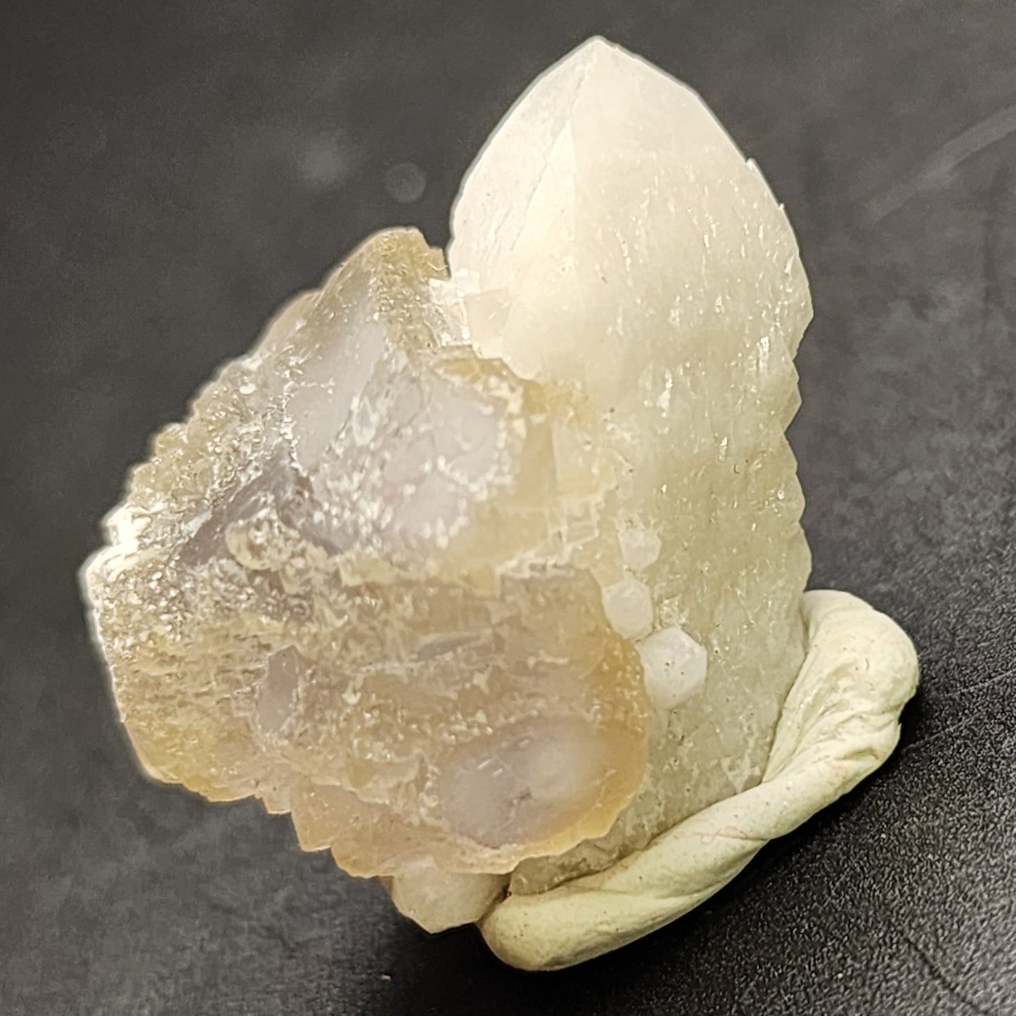 A close-up image of a tan and white crystal specimen, likely from the AUCTION Fluorite collection by The Crystalary, sourced from Huanggang, Hexigten Banner, Chifeng City, Inner Mongolia. The crystal is positioned upright on a dark surface and supported by a beige, putty-like substance. One side of the specimen showcases a rough, jagged texture while the opposite side displays a smooth, pointed formation.