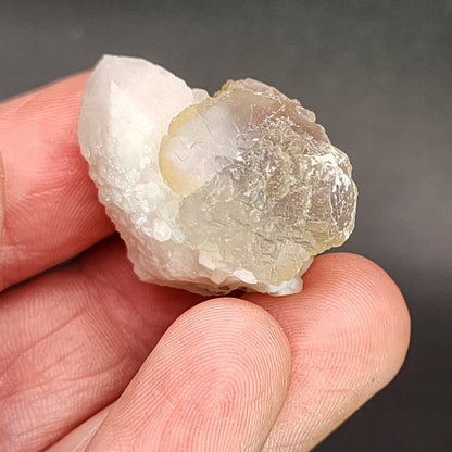 A hand holding a translucent crystal cluster against a dark background. The cluster, an AUCTION Fluorite from Huanggang, Hexigten Banner, Chifeng city, Inner Mongolia by The Crystalary, consists of milky white and light brownish-yellow crystals with intricate natural formations and textures. The fingers are slightly visible framing the crystal.