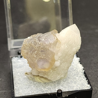 A close-up image of a mineral specimen from The Crystalary displayed on a black base with a clear protective cover. The Fluorite crystal, sourced from Huanggang, Hexigten Banner, Chifeng City in Inner Mongolia, features milky white and yellow hues with a rough, textured surface. The dark background beautifully highlights the crystalline structure.