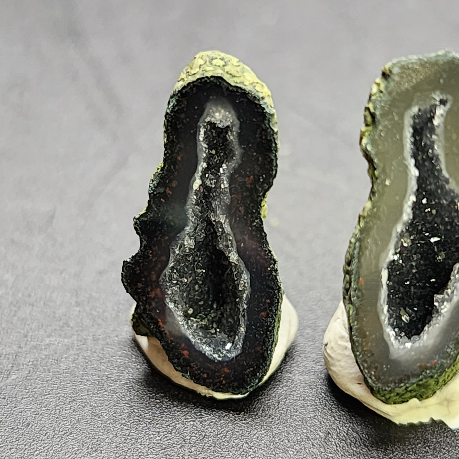 Close-up image of two small Tabasco Geode halves positioned upright. Each geode displays a rough, textured exterior and a sparkling, crystal-filled interior with a dark, somewhat reflective cavity. The geodes are set against a plain gray background. Featured Product: AUCTION- Tabasco Geodes- Tabasco, Mexico by The Crystalary.