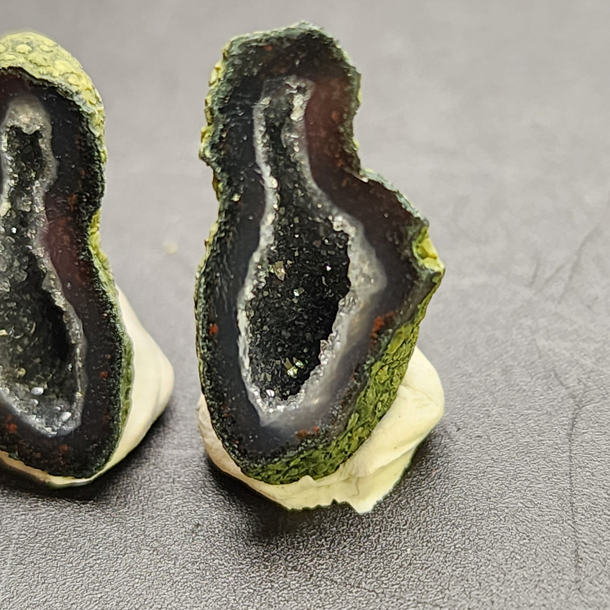 Close-up of a pair of small, sliced AUCTION- Tabasco Geodes from Tabasco, Mexico displayed upright on a gray surface. The geodes from The Crystalary feature a greenish-gray outer crust and a sparkling inner cavity with dark and light crystal formations, resembling miniature caverns.