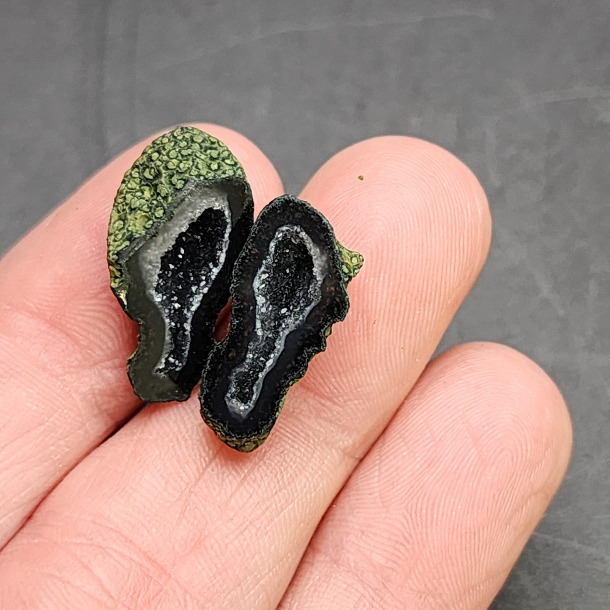 A person holding a pair of sliced AUCTION- Tabasco Geodes from The Crystalary, sourced from the Ojuela Mine in Tabasco, Mexico. The geodes showcase a dark interior filled with sparkling crystals against a dark background. Their texture is rough on the outside and glossy on the inside.