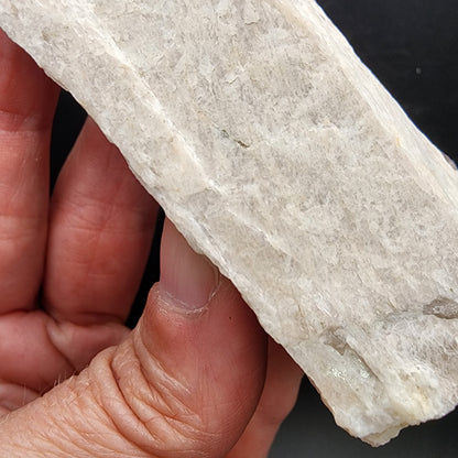 A close-up view of a hand showcasing the AUCTION Raw Moonstone from The Crystalary, featuring its rough surface and light color. This specimen, hailing from Revelstoke, British Columbia, Canada, boasts a jagged texture that stands out against the dark background.