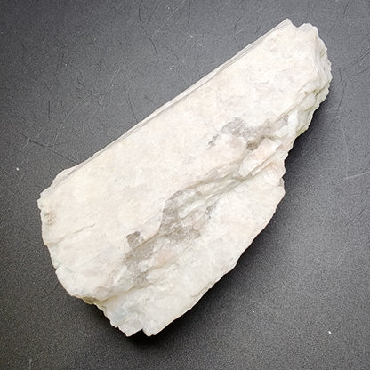 A rough, white piece of AUCTION Raw Moonstone from Revelstoke, British Columbia, Canada by The Crystalary rests on a dark, textured surface. The stone's uneven edges and slightly translucent texture highlight its natural grooves and imperfections, embodying the untouched beauty reminiscent of its origin.