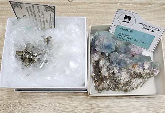 Displayed in white boxes are two mineral specimens from The Crystalary's LIVE- vineyardguy- 9/26/2024 collection. The left box showcases white quartz and metallic pyrite crystals nestled on bubble wrap, while the right box features a piece of rock adorned with blue, green, and purple Okorusu fluorite crystals, accompanied by labeled cards reading "FLUORITE" and indicating the origin from Namibia.