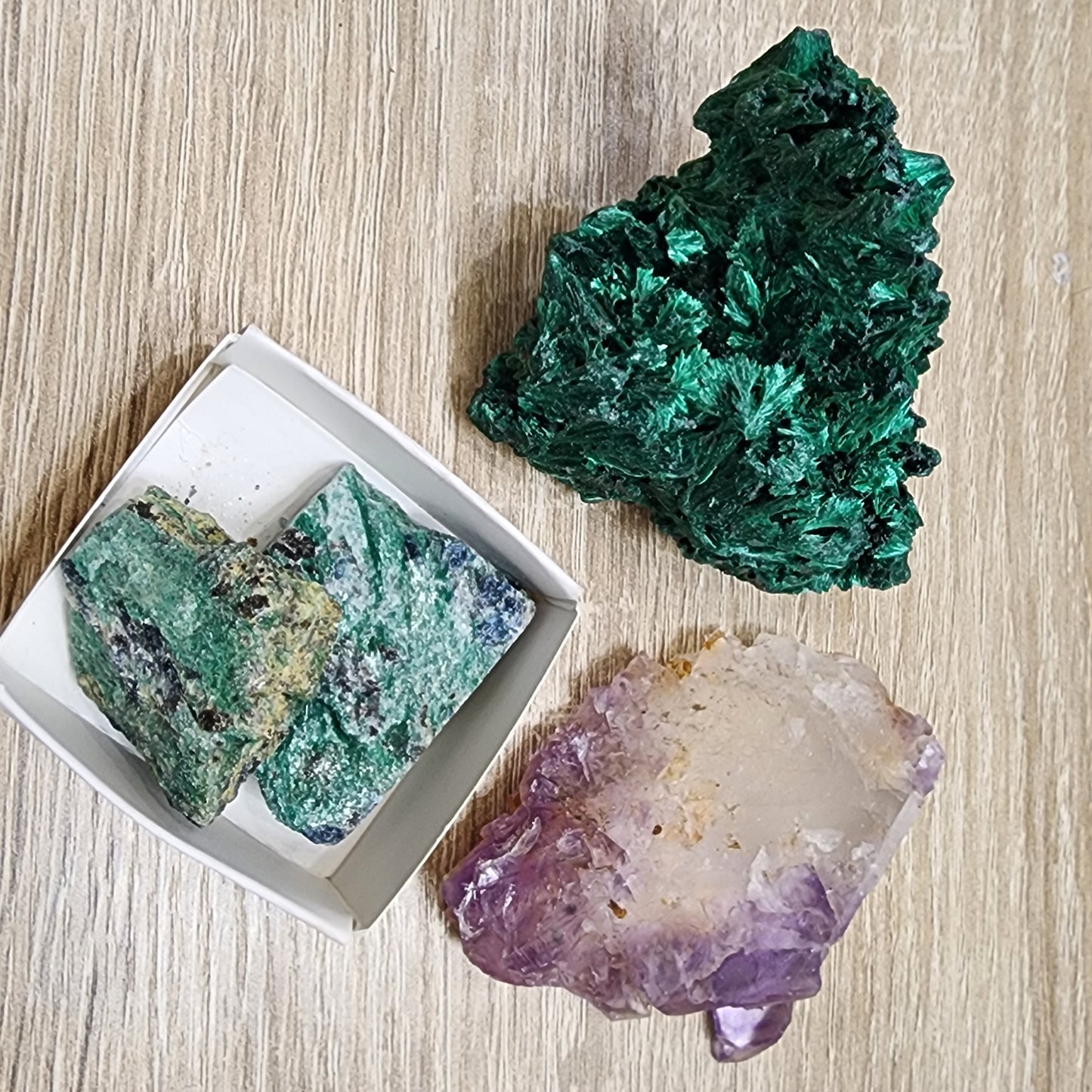 A collection of minerals and crystals displayed on a wooden surface. In the top right corner, there's a green, leafy Malachite cluster. At the bottom center is an amethyst radiating purple hues. On the left side, a rectangular box contains two rough-textured Green Mica specimens. Perfect for our upcoming The Crystalary LIVE- MindyInked- 9/26/2024 GIVEAWAY!