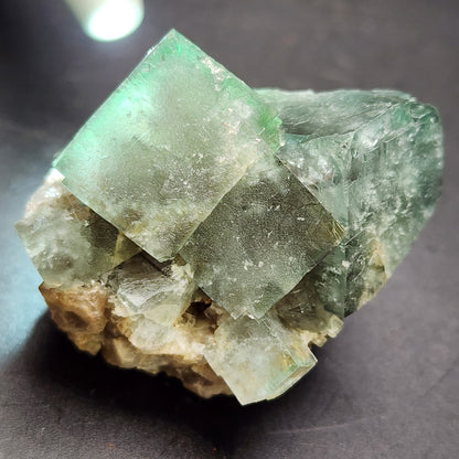 Close-up of an emerald-green fluorite crystal with a slightly translucent appearance, resting on a rough rock base. The crystal displays natural imperfections and shines under the light, highlighting its geometric structure against a dark background—reminiscent of the treasures from The Crystalary's "Fluorite - Fairy Holes Pocket," sourced from Lady Annabella Mine, Eastgate, Weardale, Co. Durham, England.