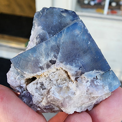 A hand holding a specimen of fluorite crystals from the Fairy Holes Pocket, Lady Annabella Mine, Eastgate, Weardale, Co. Durham, England. The piece, provided by The Crystalary, features distinct angular facet patterns and shines with a semi-translucent appearance against an indoor setting with blurred objects in the background.