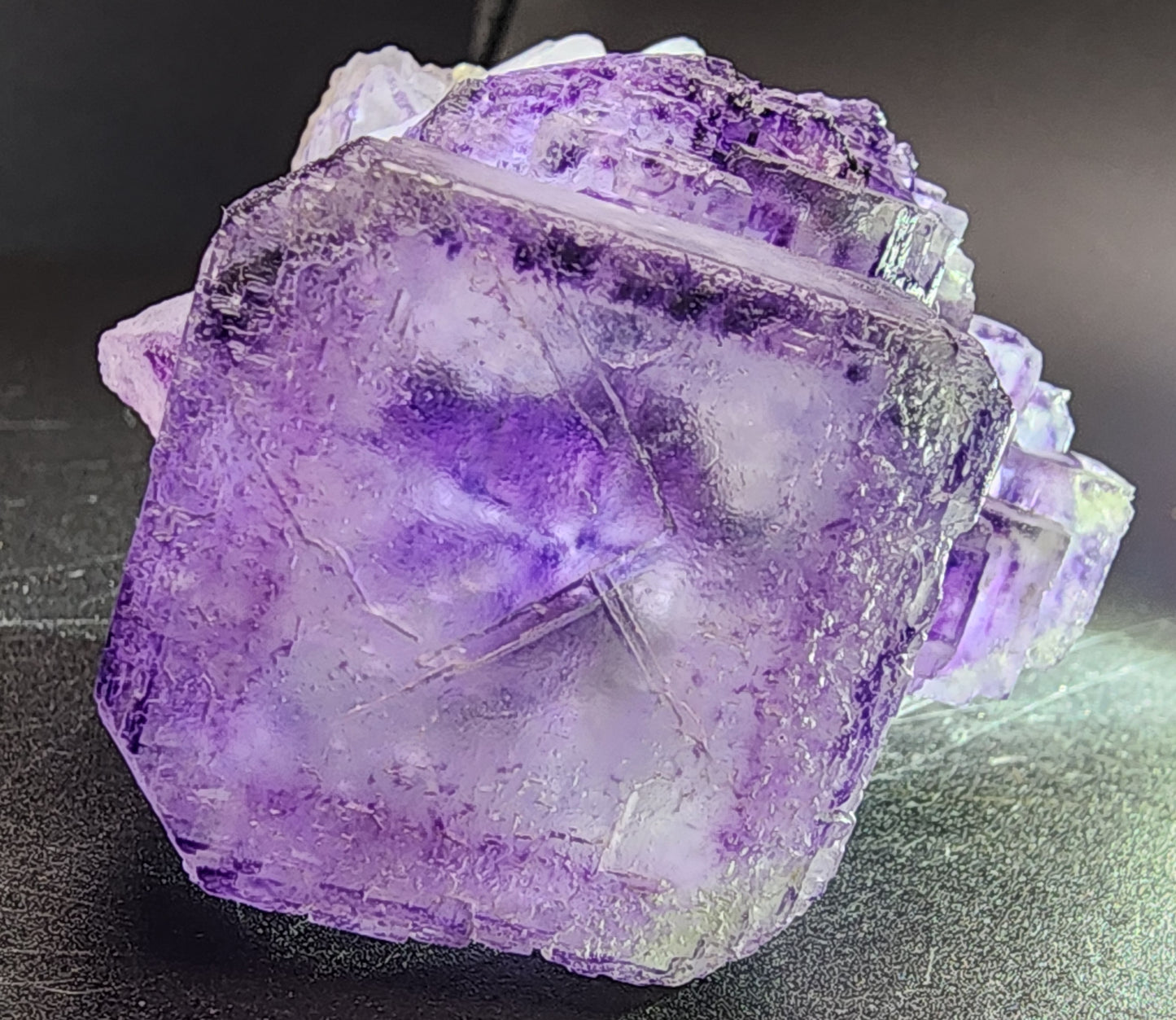 A detailed view of the Fluorite cube from Larkin's Quarry, available as sku3000 under The Crystalary brand, highlights its natural geometric formation with a textured surface against a dark background. This beveled cube exhibits vibrant colors in diverse shades of purple along with translucent sections that reveal distinct phantoms within its structure.