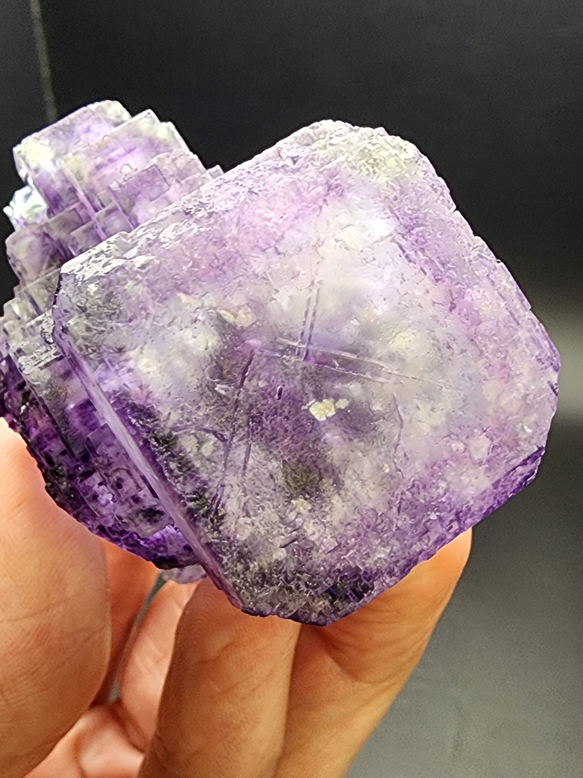 A close-up captures a hand holding The Crystalary's Fluorite from Larkin's Quarry, Connemara, Ireland (sku3000), against a dark background. The large geometric crystal displays a rough texture with visible layers showcasing shades of violet and lavender throughout, and its vibrant color accentuates the dazzling clear phantoms within.