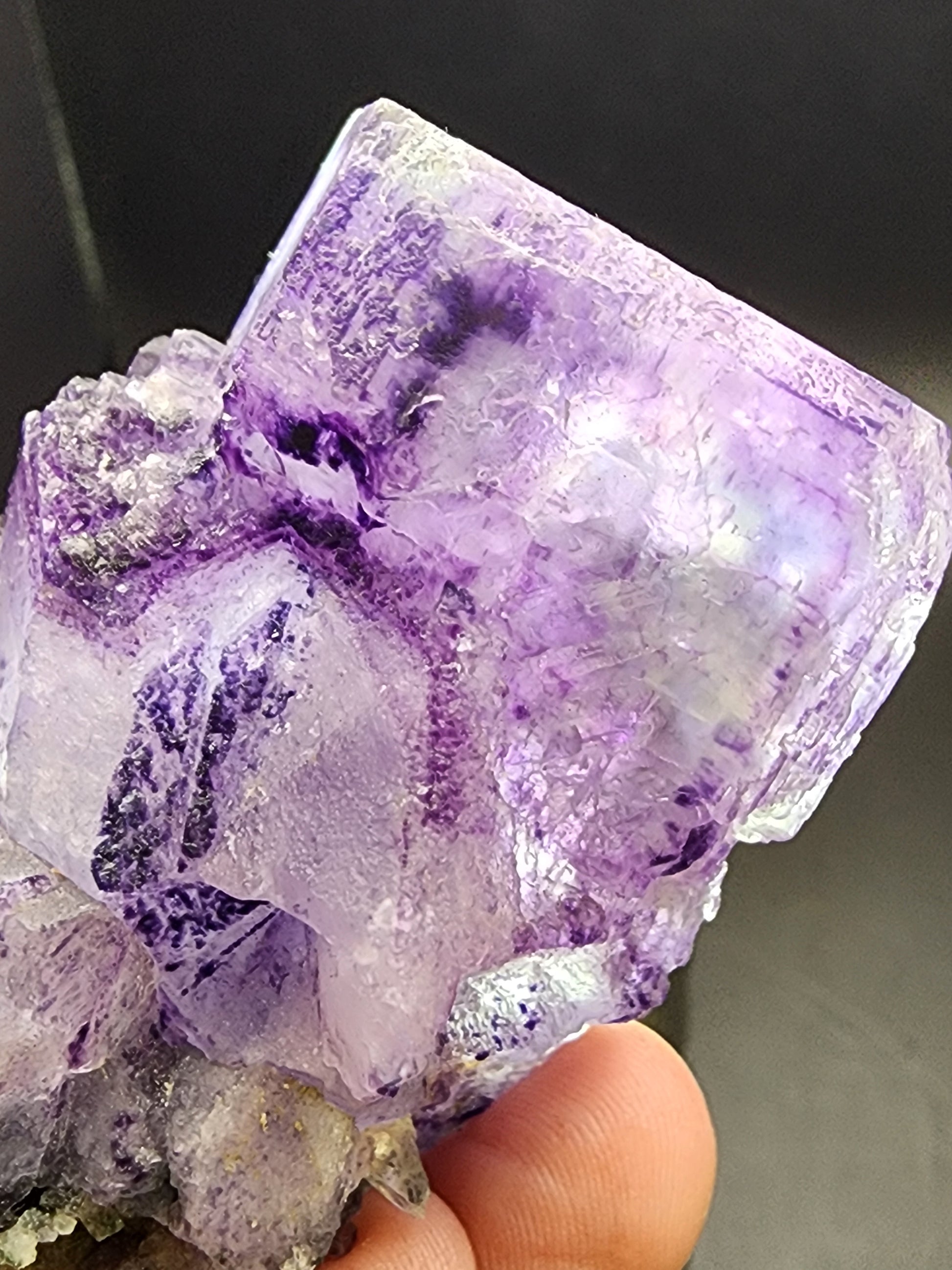 A detailed close-up image features a hand holding The Crystalary's Fluorite from Larkin's Quarry, Shannapheasteen, Costelloe, Connemara, Galway County, Connacht, Ireland (sku3000). This mineral specimen is purple and translucent with visible phantoms in its cubic formations. Its rough and jagged surfaces display hues of purple and white, with certain areas appearing shiny and reflective.