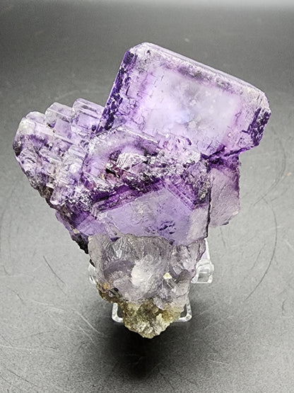 The Fluorite from The Crystalary, sourced from Larkin's Quarry in Connemara, Galway County, Ireland (sku3000), features purple cubic crystals with beveled edges and a clear geometric structure. These crystals rest on a small rocky base set against a dark background, showcasing translucency and striking variations in color and texture.