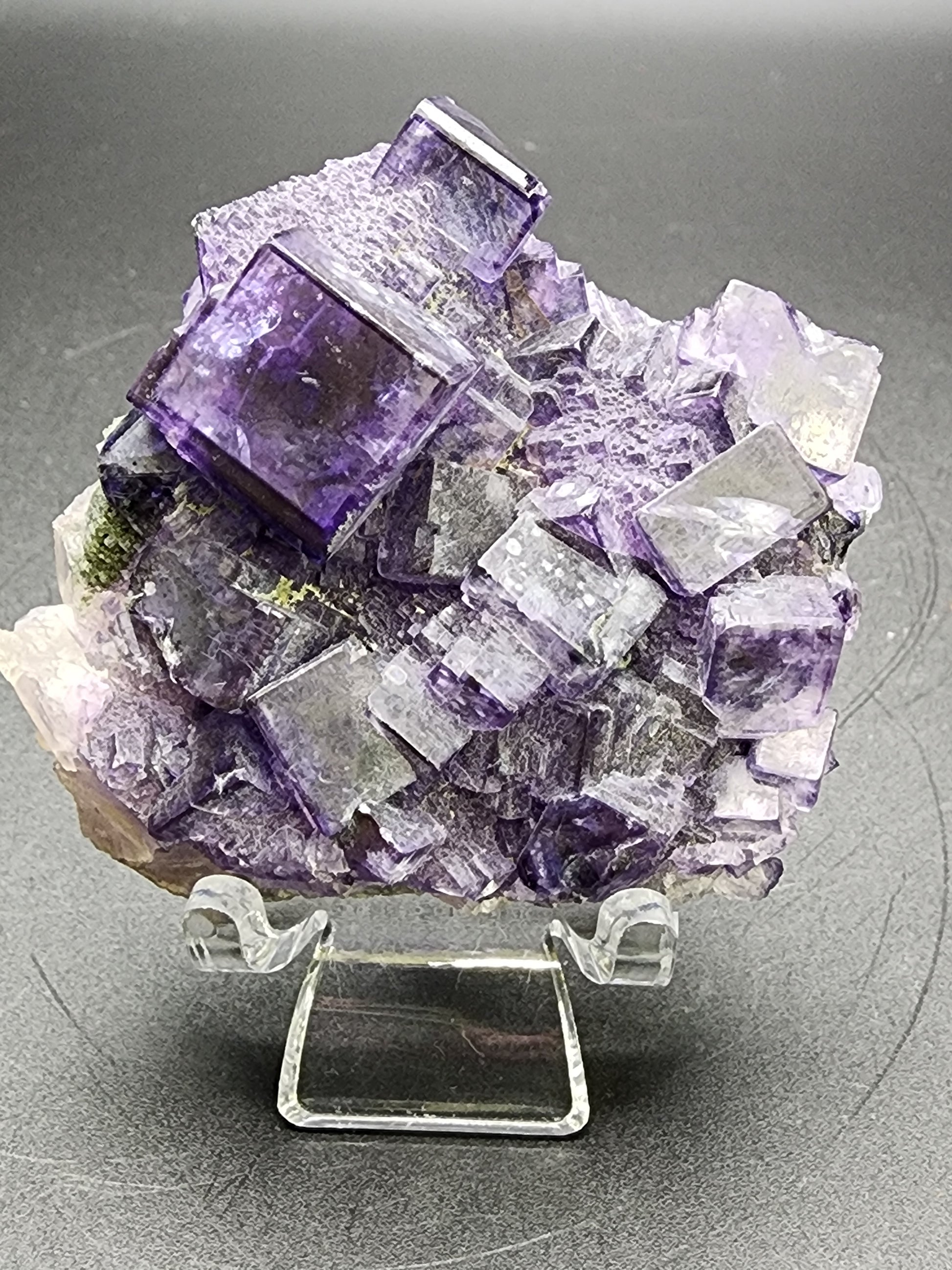 A medium cluster of purple and clear crystals with high luster cubes sits on a clear display stand, reminiscent of the treasures from The Crystalary's Fluorite collection at Larkin's Quarry, against a dark background.