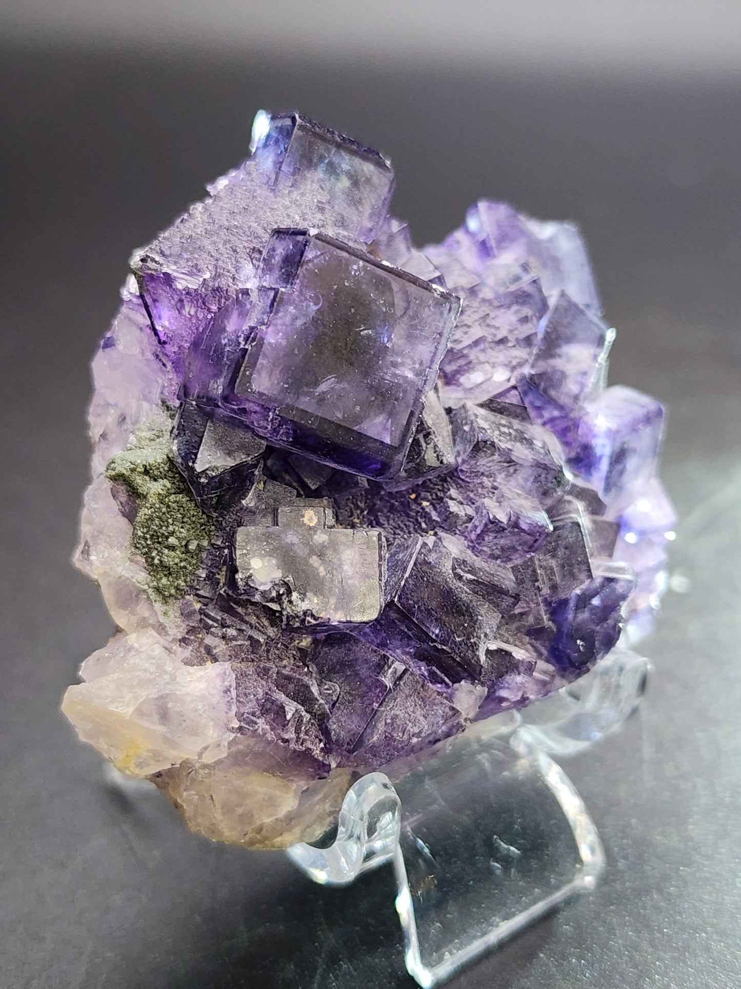 These medium-sized, high-luster cubes of purple fluorite crystals from Larkin's Quarry in Galway County are presented on a reflective surface. The translucent gems from The Crystalary display a range of purple shades with green inclusions, contrasted against a soft gray gradient.