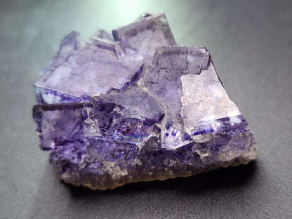 A detailed image features a raw Fluorite crystal from The Crystalary, sourced from Larkin's Quarry in Shannapheasteen, Costelloe, Connemara, Galway County, Connacht, Ireland (sku3001). The deep purple crystal exhibits sharp, angular edges and a translucent layered structure on a dark background, reminiscent of stained glass.