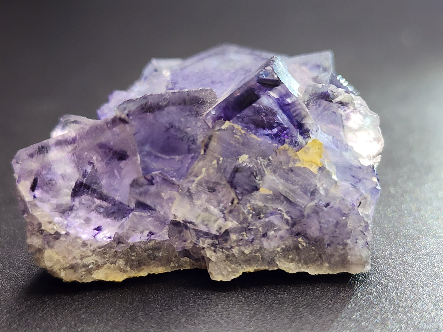 A close-up of The Crystalary's Fluorite from Larkin's Quarry, SKU: 3001, set on a black surface, highlighting its intricate geometric forms and layered texture. The lighting enhances the crystal's stained glass effect, displaying various shades of purple with hints of yellowish tint.